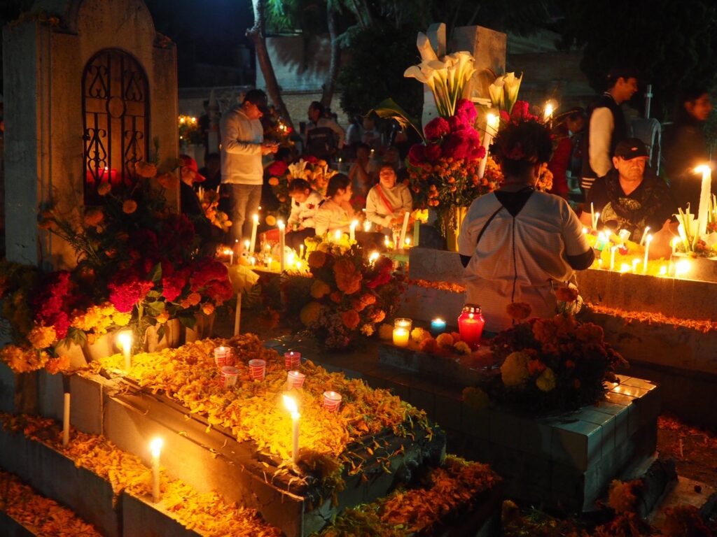 A nighttime scene at a Mexican cemetery during Día de Muertos, showing families gathered around graves decorated with bright orange marigolds (cempasúchil) and hundreds of lit candles, creating a warm and spiritual glow.