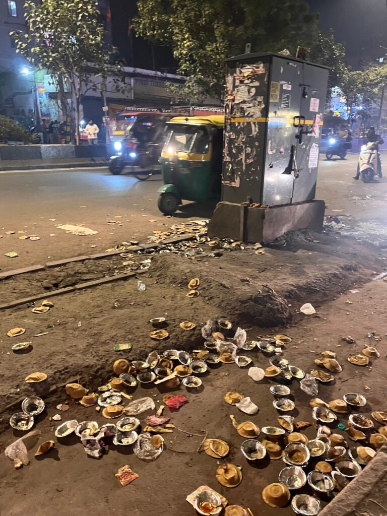 A night street scene in India showing a green and yellow auto-rickshaw parked next to a utility box, with a large amount of discarded disposable food bowls scattered on the dirt ground.