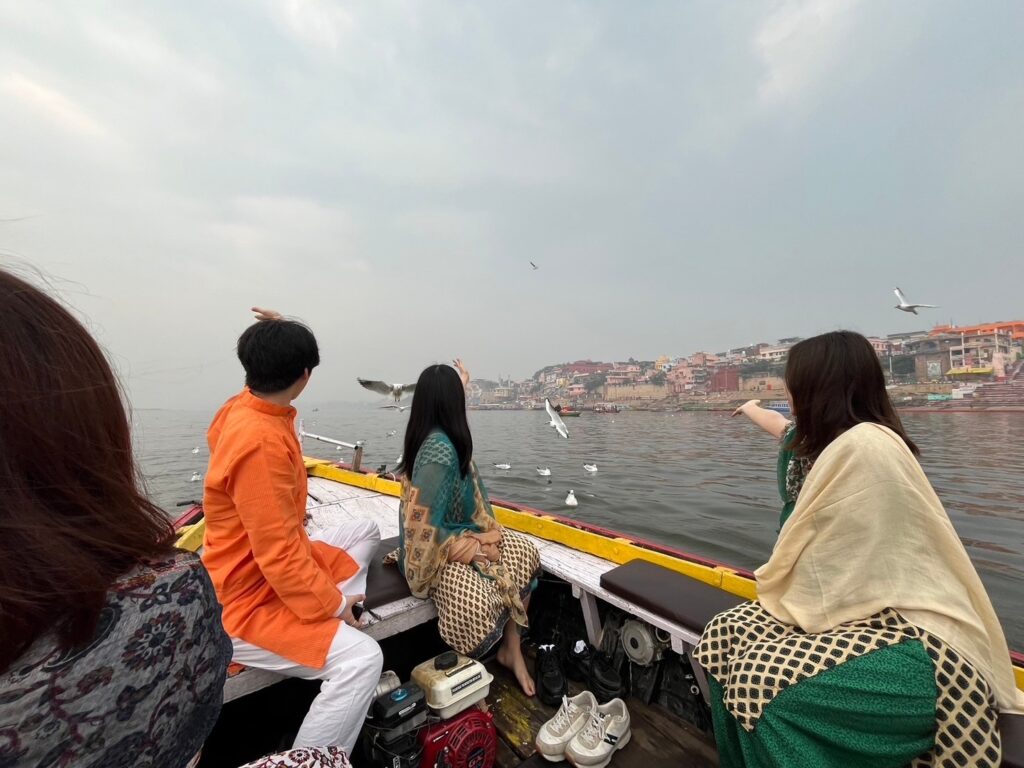 Three travelers in traditional Indian clothing sitting on a boat in the Ganges River, pointing at white seagulls flying over the water in Varanasi.