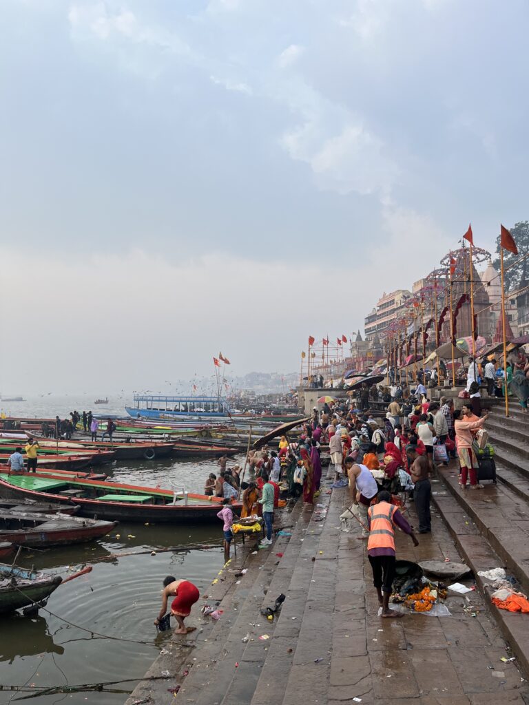 A crowded scene at the ghats of Varanasi along the Ganges River under a cloudy sky, featuring numerous wooden boats on the water, pilgrims bathing, and people gathering on the stone steps with temples in the background.