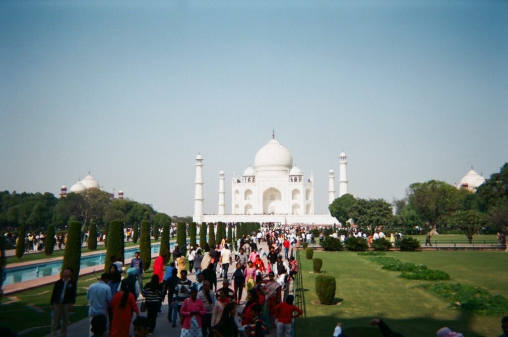 The white marble Taj Mahal mausoleum in Agra, India, with its minarets and dome, viewed from the long, manicured gardens and reflecting pools crowded with tourists.
