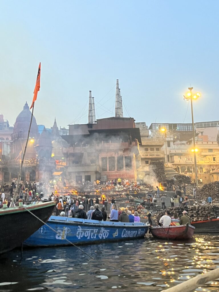 A view of the Manikarnika cremation ghat in Varanasi from a boat on the Ganges River, showing smoke rising from funeral pyres, stacks of wood, and people observing the ancient rituals at dusk.