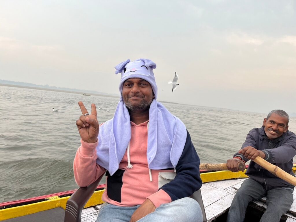 A friendly Indian man wearing a whimsical purple animal hat with ears, smiling and making a peace sign on a boat in Varanasi. An elderly boatman is rowing in the background on the Ganges River.