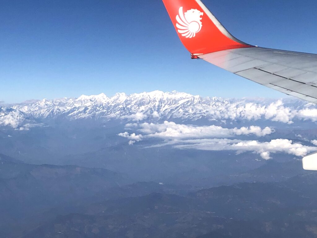 Aerial view of the snow-capped Himalayan mountain range under a clear blue sky, taken from an airplane window with the red and white wing of a Lion Air aircraft in the foreground.