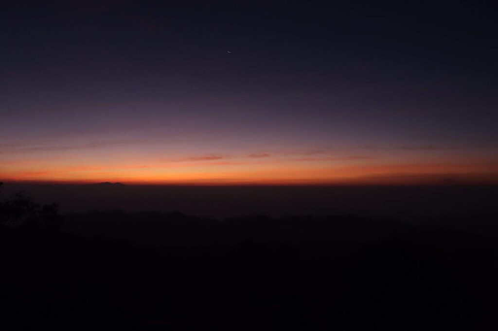 A beautiful sunrise or sunset over a mountain range, featuring a deep purple sky transitioning into a bright orange horizon with a faint crescent moon visible above the clouds.