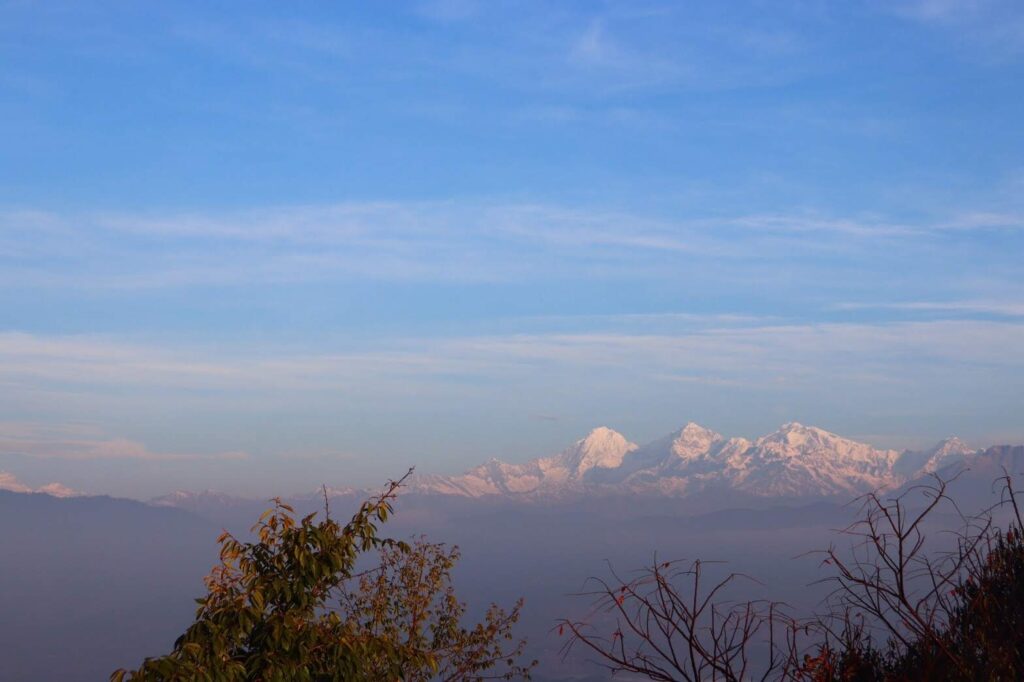 Distant snow-covered mountain peaks under a clear blue sky, partially obscured by morning mist and foreground trees.