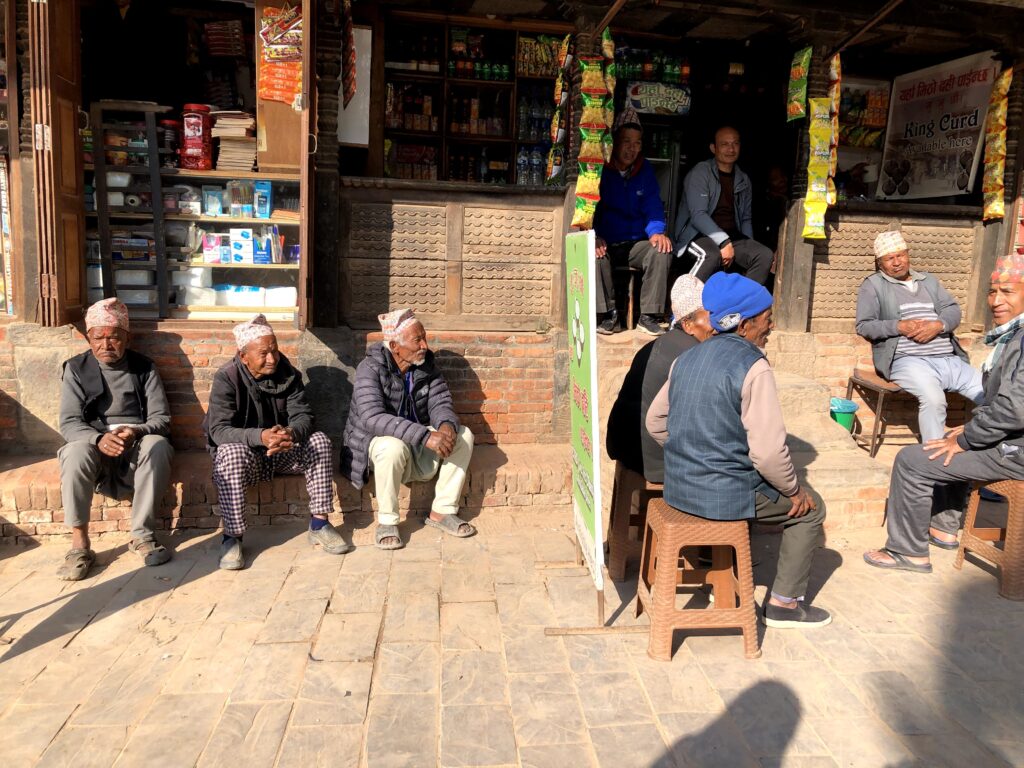 Local men sitting and socializing in front of a traditional Nepali shop in Bhaktapur, with a green "King Curd" (Juju Dhau) advertisement board and snacks displayed in the open storefront.