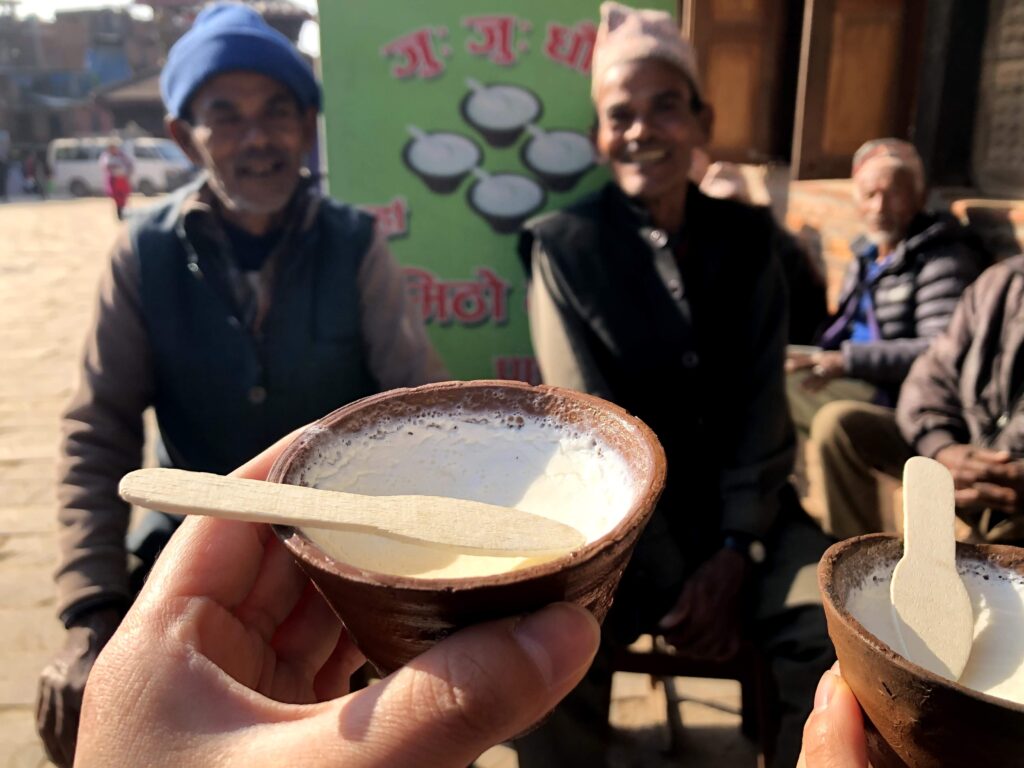A close-up of a hand holding a traditional clay bowl of creamy King Curd (Juju Dhau) with a wooden spoon, with smiling local men and a green advertisement board in the blurred background.