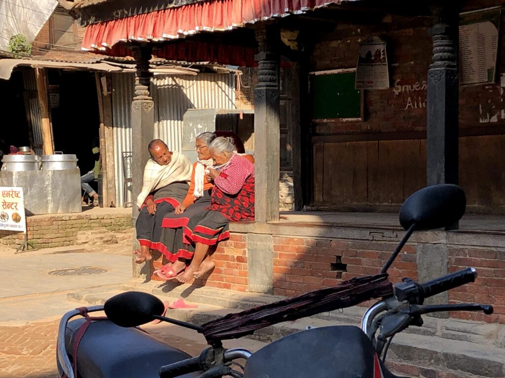 Three elderly Nepali women in traditional dress sitting and talking on the brick ledge of a historic wooden building in Bhaktapur, enjoying the afternoon sun.