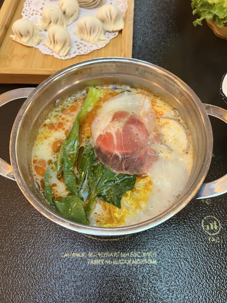 A stainless steel pot of Mongolian hot pot with meat, green vegetables, and a milky broth, with a tray of raw dumplings in the background.