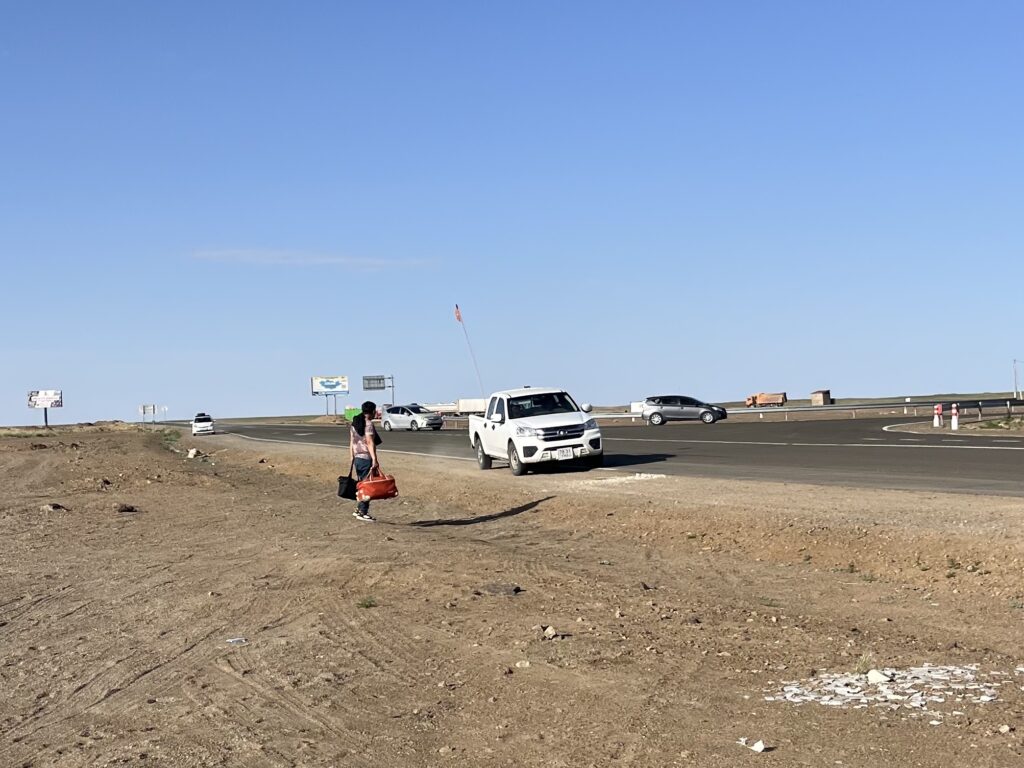 A lone traveler carrying bags walking along the dusty shoulder of a paved highway in the Mongolian steppe, with a white pickup truck and other cars driving past under a clear blue sky.