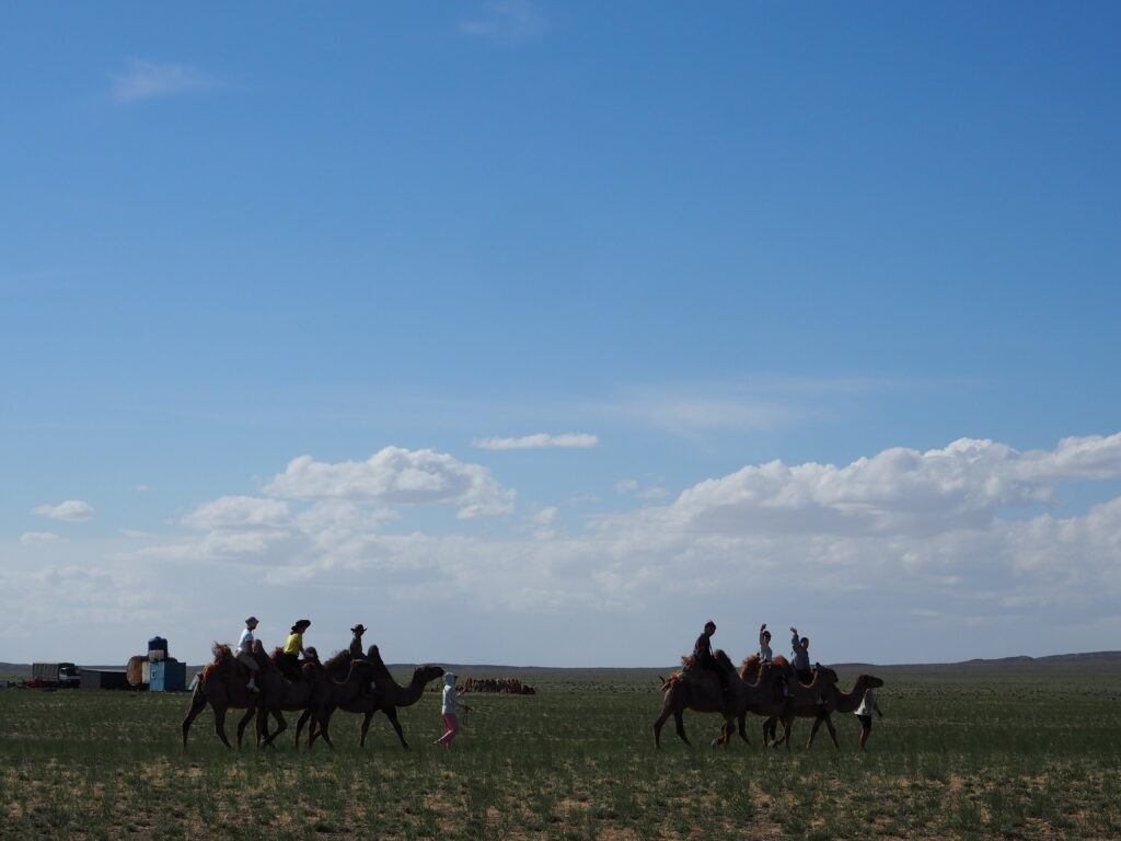 A wide-angle view of a herd of goats and sheep grazing on the dry Mongolian plains near a small, colorful yellow and blue roadside building under a dramatic cloudy sky.