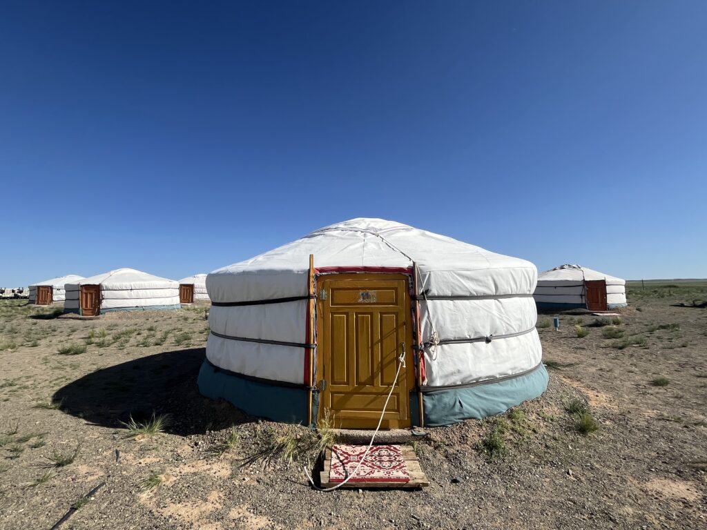 A close-up of a traditional Mongolian Ger (Yurt) with a wooden orange door and a patterned rug at the entrance, with other white Gers scattered across the dry landscape.