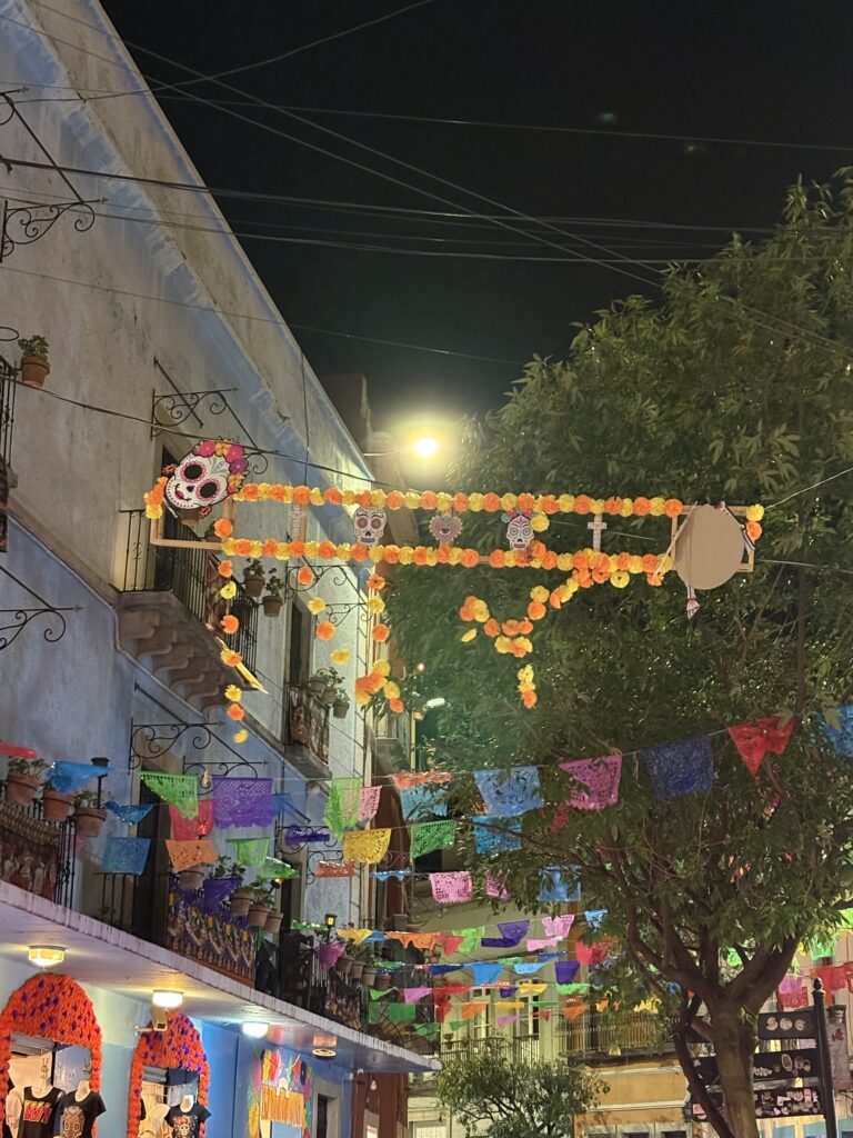 A vibrant night street scene decorated with colorful papel picado banners and Día de Muertos ornaments, featuring sugar skull decorations and orange marigold garlands hanging between historic buildings.